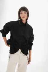 Camisa blanca de mangas largas con cuello clásico y cierre con botones. Presenta detalles de fruncido vertical en la parte delantera y cordones ajustables en los laterales. Corte holgado.