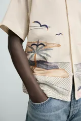 Camisa de manga corta y calce relajado en color beige, con cuello bowling y cierre frontal de botones. Presenta un diseño bordado en la parte inferior con motivos de paisaje tropical que incluyen palmeras, sol y aves.