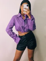 Camisa cropped negra con cuello clásico, mangas largas con puños abotonados y cierre frontal con botones. Presenta detalle de tachas plateadas en el cuello.