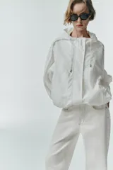 Chaqueta blanca de cuello redondo con capucha ajustable mediante cordones elásticos. Presenta manga larga con puños elásticos, bolsillos delanteros y bajo asimétrico ajustable con cordones y stoppers. El cierre frontal combina cremallera y botones ocultos bajo una solapa.