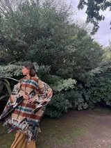 Poncho estilo navajo confeccionado en mezcla de lana y algodón, con estampado geométrico multicolor en tonos beige, marrón, negro, turquesa y blanco, y terminación de flecos en el ruedo.
