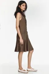 Vestido midi camisero color marrón, sin mangas, con cuello solapa y cierre frontal de botones. Presenta bolsillos delanteros con solapa y diseño con pliegues en el bajo.