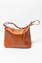 Bolso de hombro tipo hobo, confeccionado en cuero texturizado color marrón. Presenta un diseño minimalista con cierre superior, correa ajustable y detalle de tiras decorativas en el lateral.