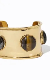 Brazalete rígido de metal dorado de banda ancha, con abertura frontal y piedras redondeadas con efecto veteado incrustadas.