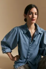 Camisa de denim azul con cuello camisero, cierre frontal de botones y mangas largas abullonadas con hombros fruncidos.