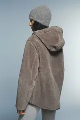 Abrigo de tejido suave efecto peluche en color marrón, con capucha ajustable mediante cordones, cierre frontal de cremallera y bolsillos laterales insertados. Presenta ribetes en los bordes y cuello elevado.