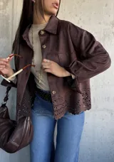 Chaqueta de gamuza color chocolate, con corte recto, cuello clásico y cierre frontal con botones. Presenta un diseño calado decorativo en el ruedo y bolsillos frontales tipo parche.