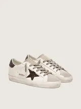 Zapatillas Golden Goose modelo Super-Star, color blanco con detalles en marrón con glitter. Confeccionadas en cuero vacuno, con estrella lateral y contrafuerte de talón brillantes, punta y lengüeta de gamuza, y efecto envejecido en la suela.
