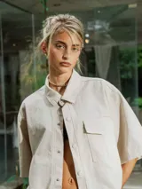 Camisa de manga corta color crudo, confeccionada en gabardina, con cuello camisero y botones frontales. Presenta un bolsillo de parche en el pecho y bordado en la espalda.