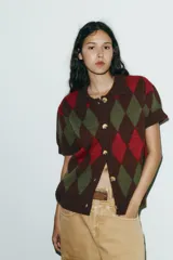 Chaqueta de punto de manga corta con cuello polo y cierre frontal de botones. Presenta un patrón de rombos (argyle) en tonos marrón oscuro, verde oliva y rojo.