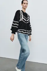 Blusa negra de manga larga con cuello redondo y cierre frontal de botones. Presenta amplios detalles de bordados y encajes blancos en el canesú frontal, mangas y ruedo inferior.