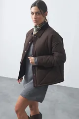 Campera tipo barn capitoneada de color marrón, con silueta corta y relajada. Presenta cuello de pana, cierre frontal con broches metálicos y bolsillos laterales funcionales.