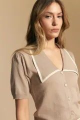 Top de tejido color beige claro, manga corta, escote en V con cierre frontal de botones y ribetes negros en el cuello y el borde del escote.