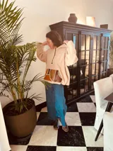 Sweater beige de corderito con capucha y bolsillo frontal de cuero en tonos marrón y ocre. Presenta tajos laterales y cierre parcial con cremallera en el cuello.