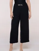 Pantalón negro de calce relajado y caída fluida, confeccionado en poliéster con textura de crepé. Tiene cintura alta elastizada estilo pull on con un aplique de cadena metálica decorativa, y pierna de corte ancho tipo wide leg corto por arriba del tobillo.