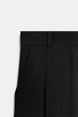 Pantalón negro de tiro alto, con trabillas, bolsillos laterales y de vivo en la espalda. Presenta pliegues en el delantero y cierre frontal con botón.