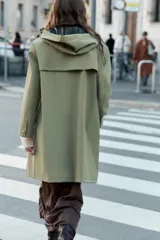 Chubasquero de acabado engomado en color verde militar, con capucha ajustable mediante cordones, cierre frontal con cremallera y botones a presión ocultos, y bolsillos laterales con solapa.