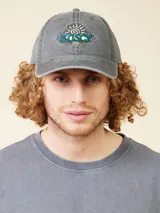 Gorro de gabardina gris oscuro con visera curva, ajustable en la parte trasera con hebilla metálica. Presenta bordado frontal con el logo de la marca y el año 1985.