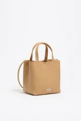Bolso pequeño de estructura rígida color beige, confeccionado en piel vacuna. Presenta doble asa de mano, correa bandolera ajustable, cierre de botón de presión y bolsillo interior con cremallera. Logo metálico de Bimba y Lola en el frente.