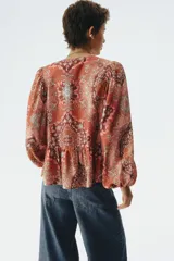 Blusa de viscosa con estampado paisley en tonos rojizos, cuello redondo con escote en V y tiras finas con cuentas decorativas. Cuenta con mangas globo largas terminadas en elástico fino y detalle de peplum acampanado en el ruedo.