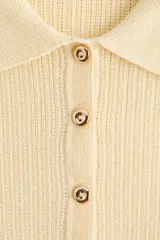 Top entallado en punto suave acanalado de color amarillo pastel, con cuello camisero, manga corta y tapeta con botones metálicos. Presenta detalles de volados en los puños y el bajo.