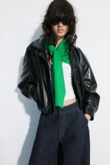Chaqueta de cuero sintético negro con cuello alto, cierre frontal de cremallera oculto por solapa con botones a presión, mangas largas y bolsillos laterales. Diseño con volumen y bajo elástico.