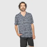 Camisa de manga corta confeccionada en viscosa, con cuello solapa y cierre frontal de botones. Presenta un estampado tropical en tonos gris y negro.