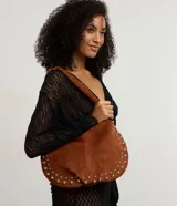 Cartera tipo baguette confeccionada en gamuza sintética color marrón. Presenta un diseño curvo con aplicaciones de tachas metálicas plateadas en los laterales, asa corta integrada y cierre mediante botón a presión.