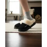 Pantuflas de gamuza negra con plataforma, forradas internamente con corderito sintético color crema. Presentan un diseño sin talón y detalle de bordado en el empeine.