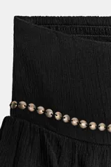 Falda pantalón corta de tiro medio, color negro, con cintura elástica en la espalda. Presenta detalle de tachas metálicas en la parte delantera y diseño de volantes a tono. Incluye forro interior.