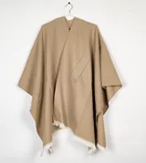 Ruana de tejido plano color beige con detalle de flecos en el ruedo y bolsillo frontal aplicado.