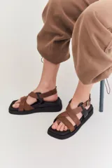 Sandalias de plataforma plana con diseño de tiras cruzadas en tonos beige y marrón. Presentan una base negra, puntera cuadrada y cierre con hebilla metálica lateral.