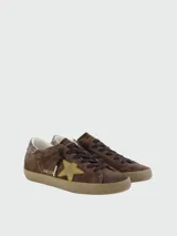 Zapatillas Golden Goose modelo Super Star, confeccionadas en gamuza color marrón con estrella lateral en gamuza dorada y detalle de glitter en el talón. Presentan cordones oscuros y suela de goma con efecto desgastado.