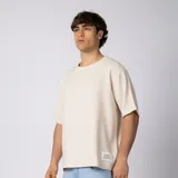 Remera oversize de lino y rayón color beige, con textura entramada, hombro caído, mangas cortas y etiqueta decorativa en el borde inferior.