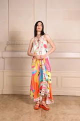 Vestido largo de seda con estampado vibrante y multicolor que incluye motivos frutales y caras sonrientes. Diseño de solera con escote halter anudado al cuello y falda amplia.