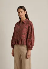 Camisa de lino con estampado floral en tonos rojizos y marrones. Presenta cuello camisero, cierre frontal con botones, mangas largas abullonadas con puños elásticos y un detalle de volado en la cintura.