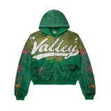 Campera con capucha y cierre frontal, de color verde con efecto desgastado. Presenta un estampado gráfico frontal con letras bordadas y aplicaciones de strass multicolor, además de detalles de llamas en las mangas y texto impreso.