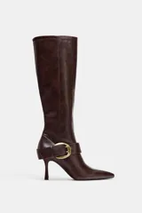 Bota de caña alta en color marrón oscuro, confeccionada en cuero con acabado liso. Presenta un diseño de punta fina, tacón alto fino y una tira decorativa en el tobillo con una hebilla metálica dorada. Incluye cierre lateral mediante cremallera.