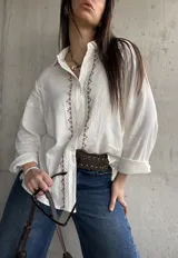 Camisa blanca de tejido liviano y calce relajado, con manga larga y detalle de puntilla y bordado en contraste a lo largo de la botonadura frontal.