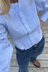 Camisa de manga larga con estampado de cuadros vichy en tonos celestes y blancos. Presenta un diseño con pliegues verticales en el frente, cuello redondo y cierre frontal con botones.