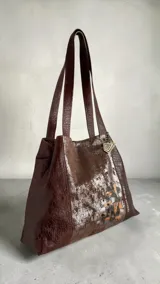 Cartera de cuero natural en color marrón jaspeado, tipo tote bag, con diseño amplio y asas dobles para el hombro. Presenta intervención artística pintada a mano en el frente con detalles metalizados y geométricos, y dije metálico colgante.