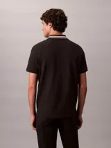 Chomba de piqué Calvin Klein color negro, con cuello clásico y ribetes a rayas blancas, cartera de dos botones y pequeño monograma CK bordado en el pecho.