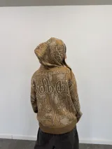 Buzo tipo hoodie con cierre frontal completo, confeccionado en tejido de punto grueso de algodón con diseño gráfico abstracto y tipográfico. Presenta capucha con bordado, puños y ruedo acanalados, y etiqueta de marca en la manga.