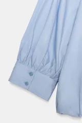 Camisa de algodón con cuello solapa, mangas abullonadas por debajo del codo y bajo acabado en globo. Presenta cierre frontal con botones ocultos.