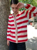 Chaqueta de punto color rojo y blanco a rayas horizontales, con ribetes y bolsillos en color rosa pálido. Presenta cierre frontal con botones dorados.