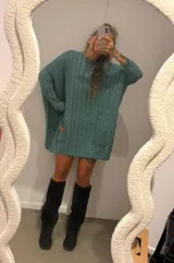 Sweater de punto color camel con diseño de trenzas verticales y acabado brillante. Presenta una silueta holgada tipo poncho, cuello redondo, mangas largas con puños ajustados y bolsillos frontales con detalle de etiqueta de cuero.