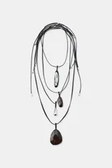 Pack de cuatro collares de cordón de piel negra con diferentes colgantes: uno con pieza metálica en forma de lágrima, otro con piedra marrón, otro con pieza ovalada blanca y negra, y otro con piedra marrón. Todos tienen cierre de lazada y acabados metálicos.