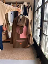 Campera de corderito marrón con capucha, diseño de apliques geométricos abstractos en tonos rosados y bordó en la parte posterior, y etiqueta frontal de la marca.