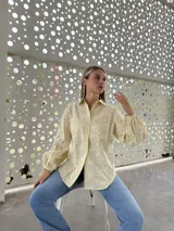 Camisa de corte holgado en color amarillo claro, confeccionada en tela con bordado suizo de motivos florales. Presenta cuello clásico, cierre frontal con botones y mangas largas abullonadas con puños elásticos.