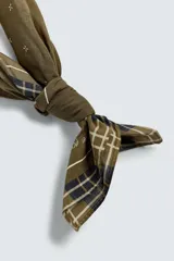 Pañuelo cuadrado de algodón color verde oliva con un patrón de pequeñas cruces blancas distribuidas por toda la superficie. El borde presenta un ribete con un patrón de cuadros en tonos oscuros (negro, azul marino) y líneas finas blancas.